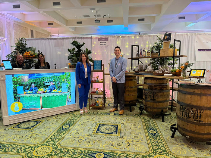 Two Executive Beverage staff beside their whiskey barrel bar and LED bar stand at a bridal showcase, with barrels and a patterned rug.