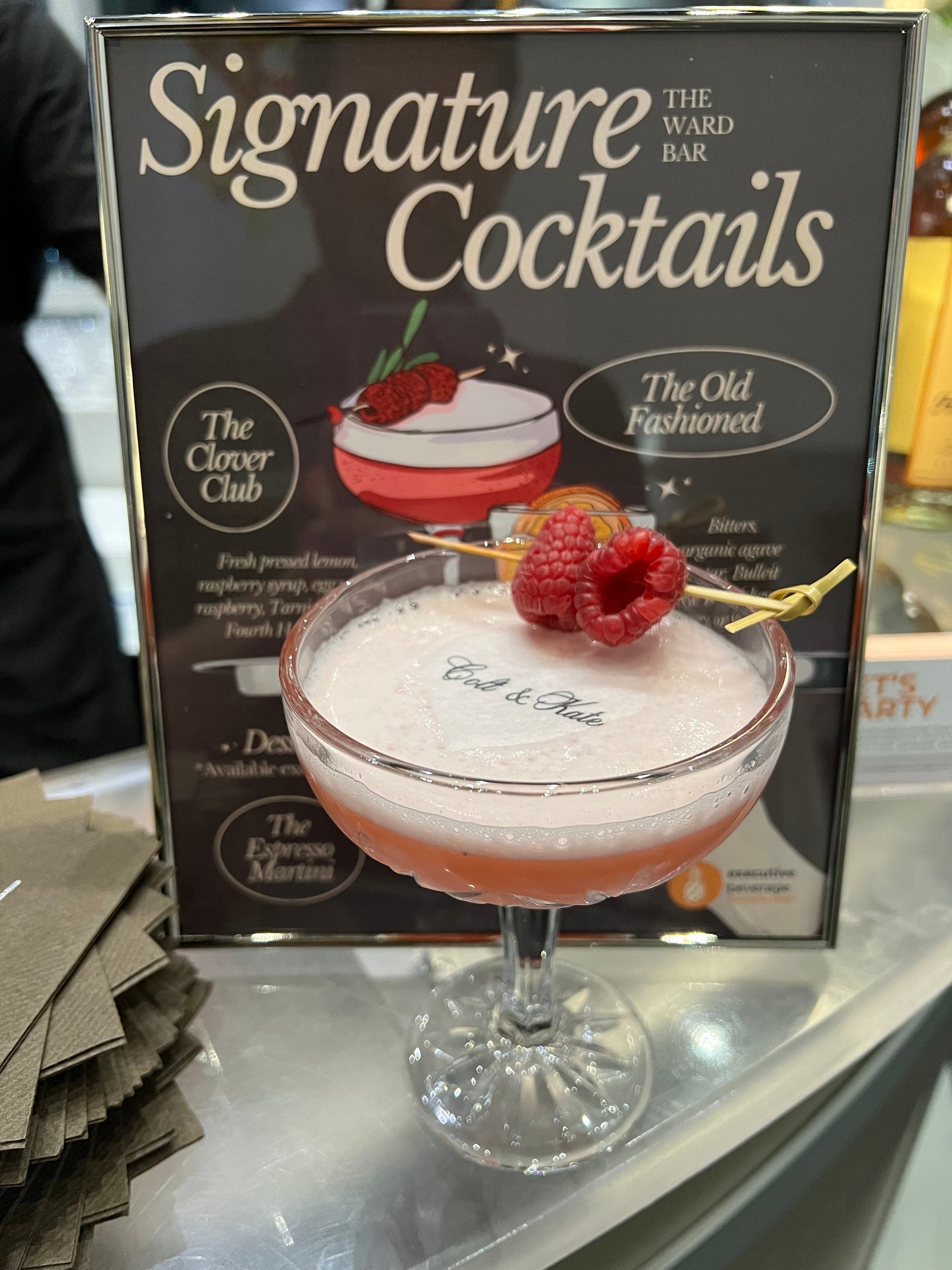 A coupe cocktail garnished with raspberries in front of a printed Signature Cocktails menu board.