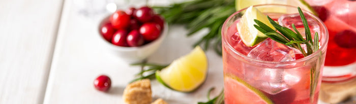 A pink infused drink garnished with rosemary and fresh cranberries beside a wedge of lemon.