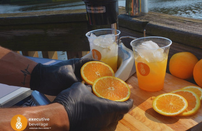 A gloved bartender preparing two orange crushes beside freshly sliced oranges on a wooden board.