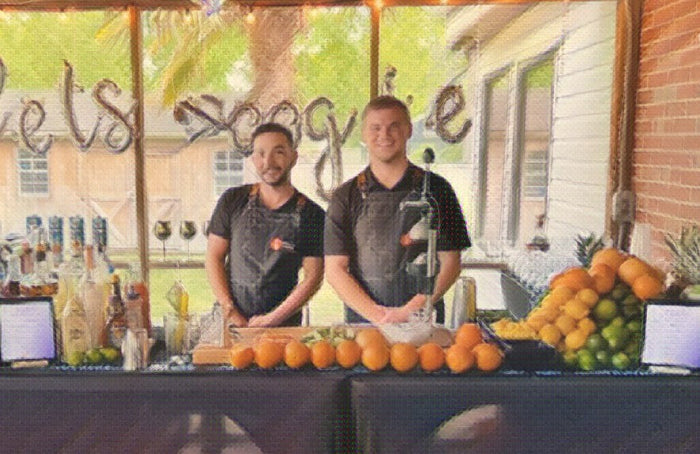 Two Executive Beverage bartenders behind a black-draped bar stacked with fresh oranges outside a venue.