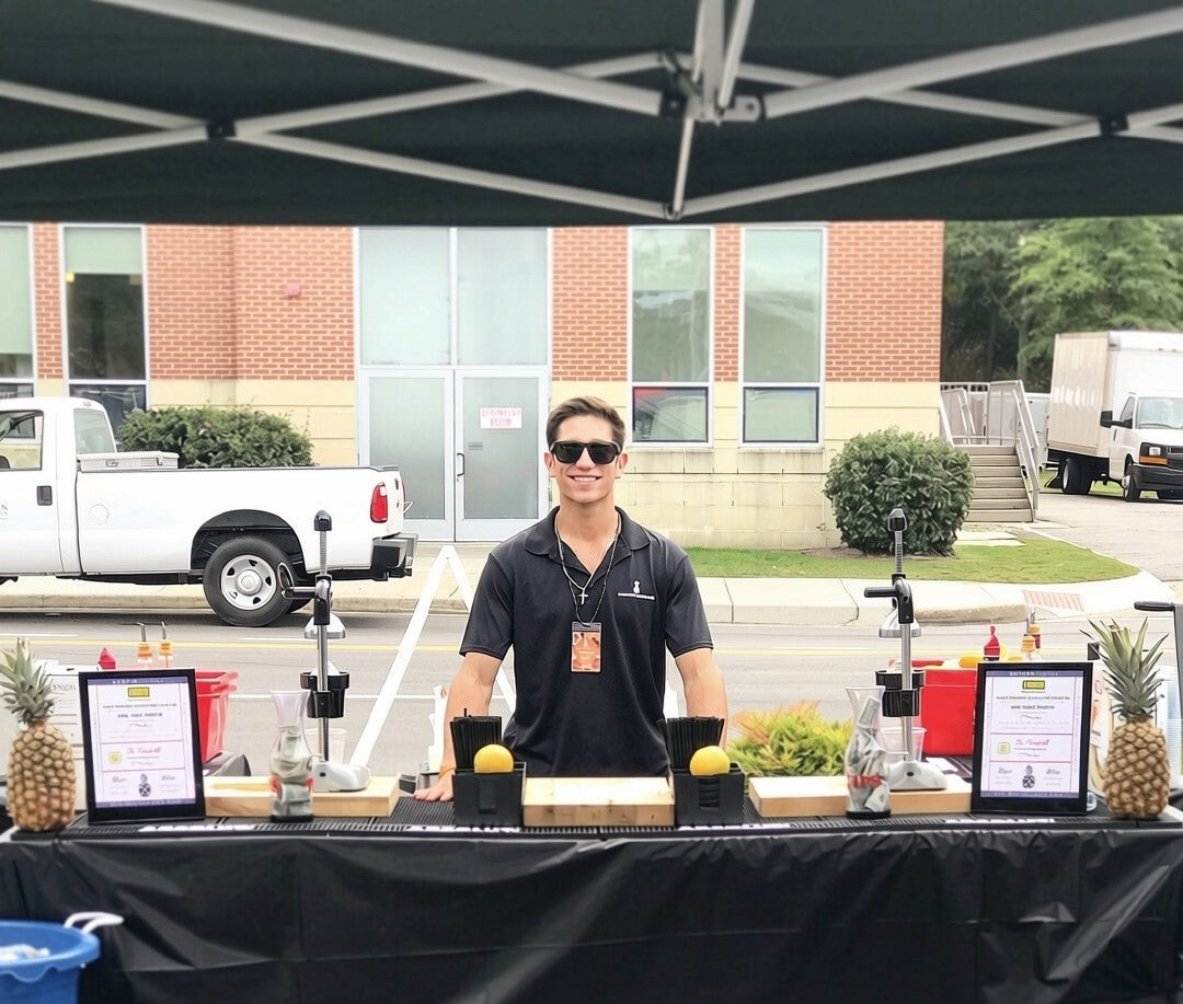 An Executive Beverage bartender behind an outdoor bar with pineapples and printed menus at a corporate site event.