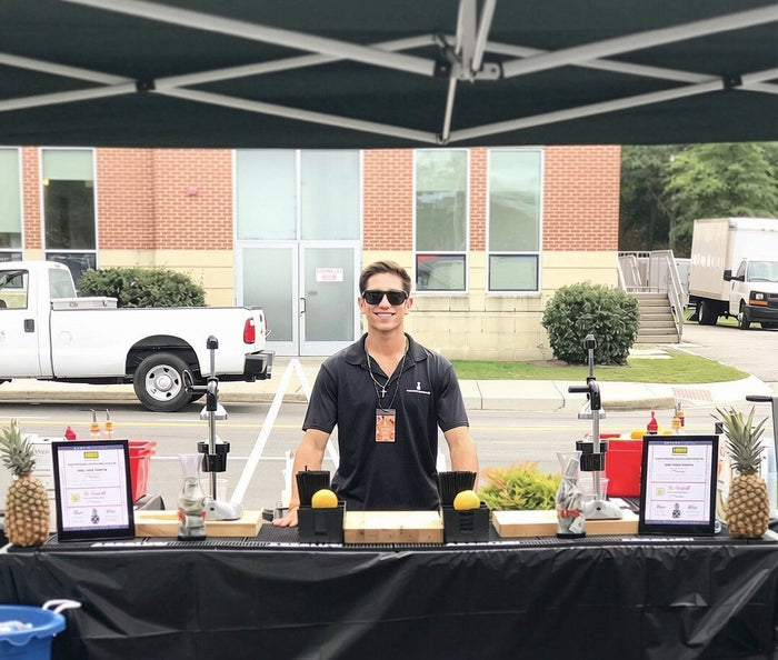 An Executive Beverage bartender behind an outdoor bar with pineapples and printed menus at a corporate site event.
