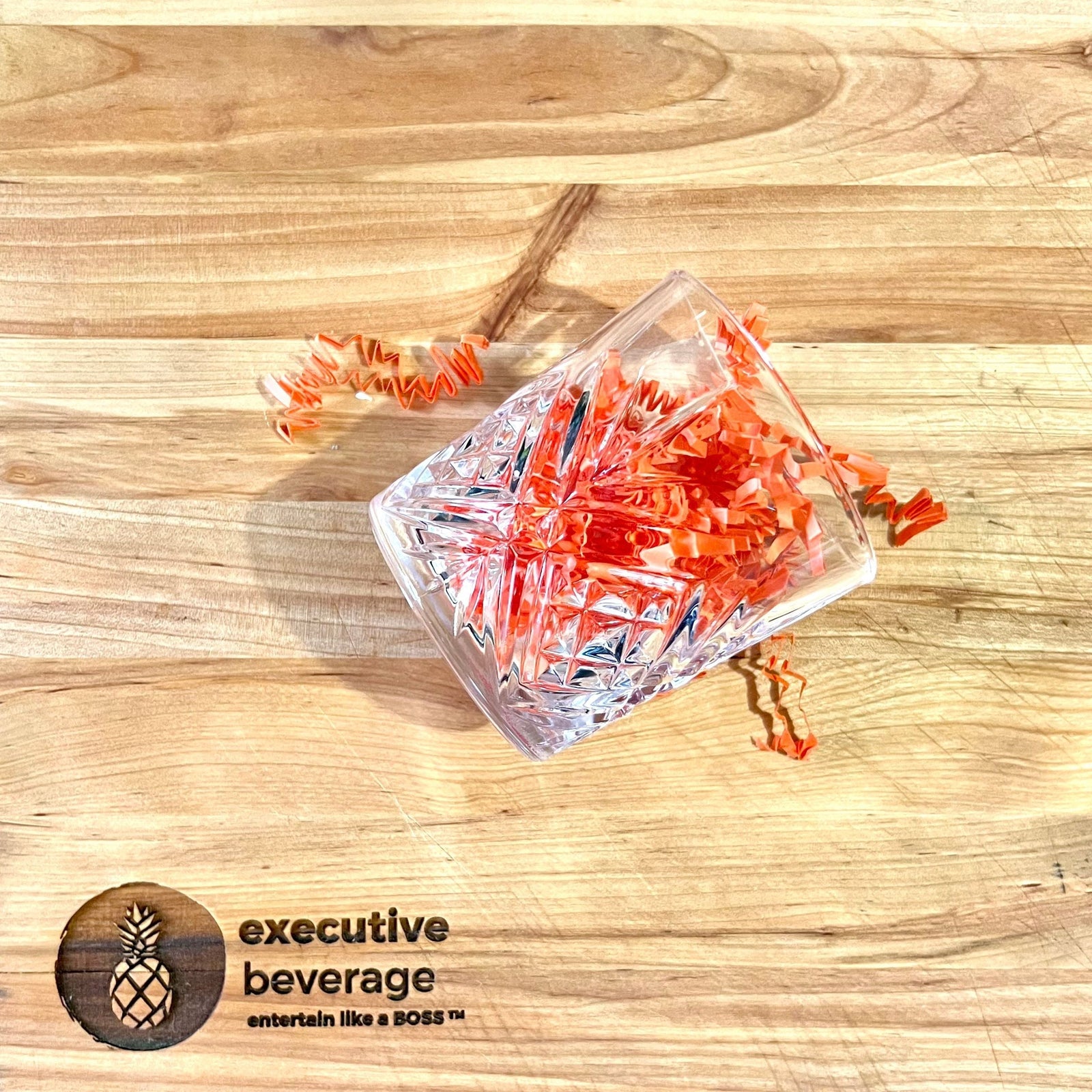 A cut-crystal rocks glass holding orange paper straws on a butcher's block, with the Executive Beverage pineapple mark.