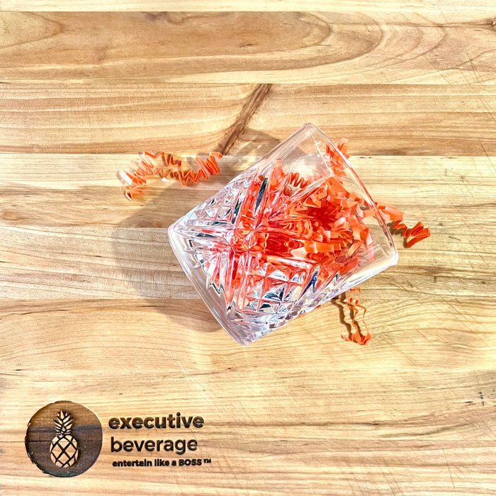 A cut-crystal rocks glass holding orange paper straws on a butcher's block, with the Executive Beverage pineapple mark.