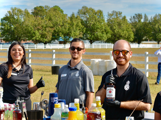 Three Executive Beverage bartenders in black polos behind an outdoor bar at a farm venue, mixers and shakers laid out.