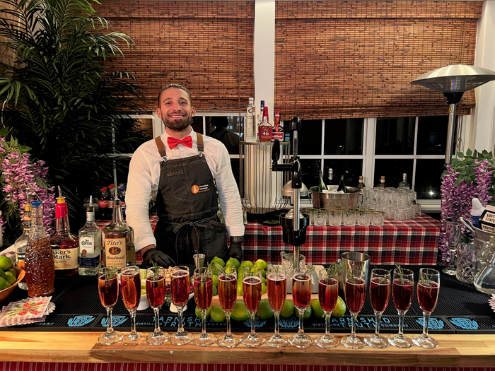 A bartender in a bow tie and apron behind a bar lined with rows of poured cocktails at an outdoor event.