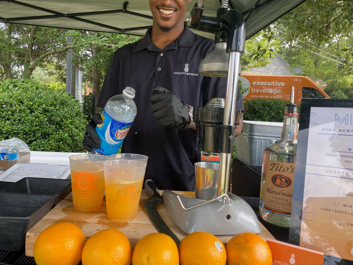 A smiling Executive Beverage bartender under a tent behind a bar of fresh-pressed orange crushes, whole oranges and spirits.