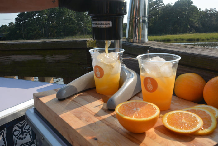 Two fresh-pressed orange crushes in branded cups on a butcher's block with halved oranges, beside the water.