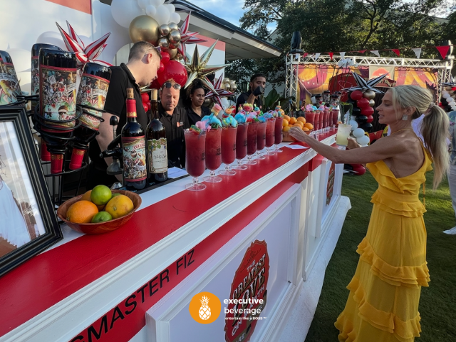 A guest in a yellow dress collecting a tall cocktail from a branded Executive Beverage bar at a busy event.