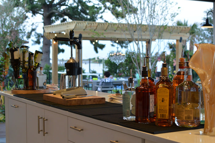 An outdoor bar under a pergola set with spirits and a cutting board beside the water.
