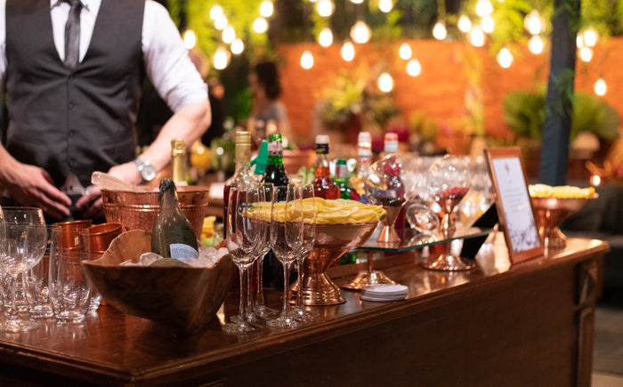 An Executive Beverage wedding bar at dusk under string lights, set with copper barware, champagne flutes, fresh citrus and a printed cocktail menu.