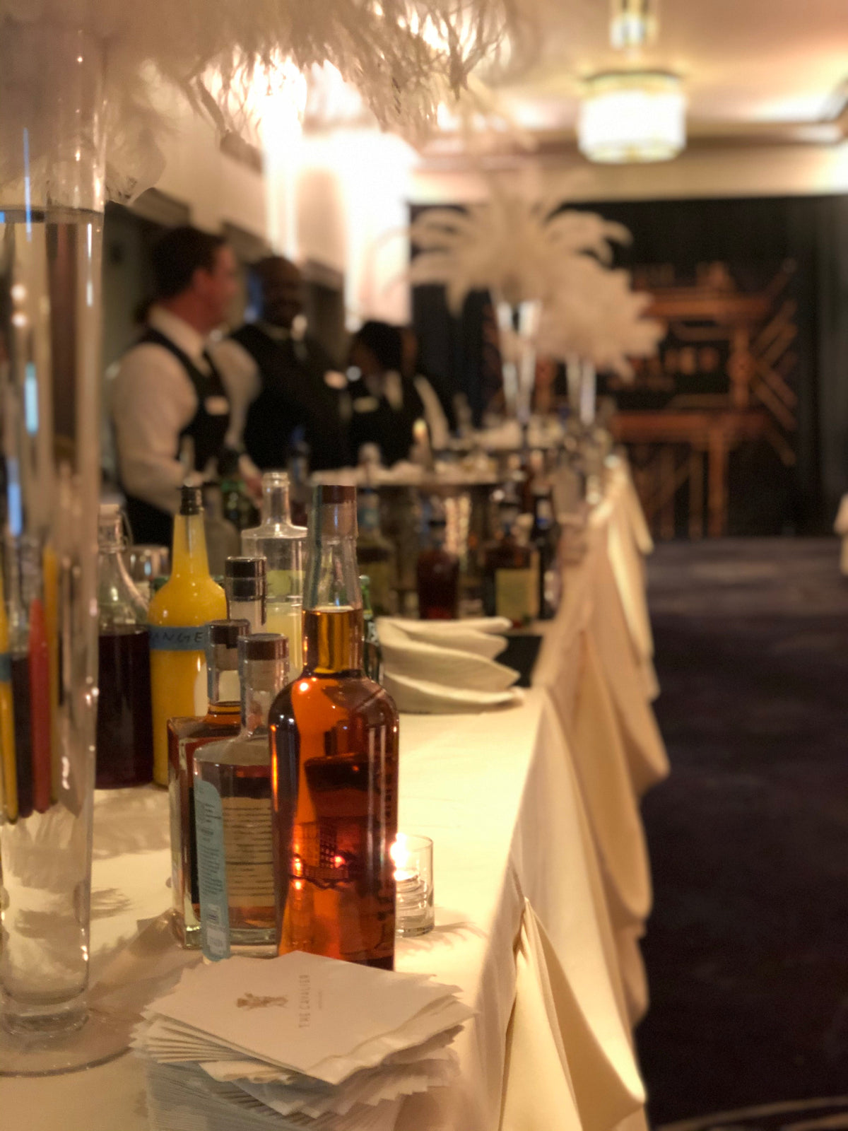 A dressed indoor bar with whiskey bottles and tall white feather centerpieces at a wedding reception.