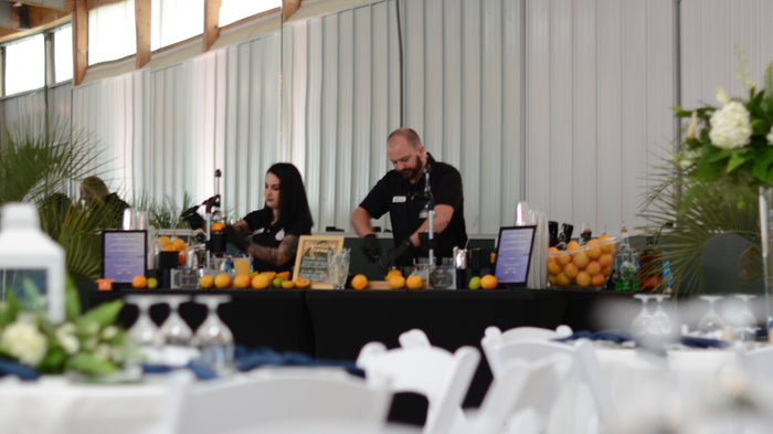 Executive Beverage staff behind a long indoor bar with digital menus, fresh oranges and white florals.