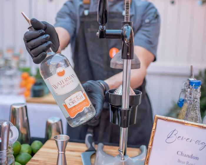 An Executive Beverage bartender in a branded apron pressing fresh juice beside Waterman Spirits crush vodka, bar tools and a printed beverage menu.