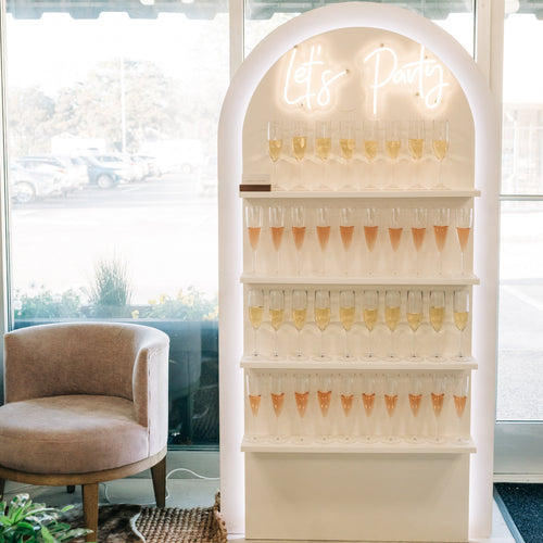 A white arched party display shelf with a glowing neon 'Let's Party' sign and multiple tiers of champagne flutes, set in a bright modern indoor space with large windows, a beige armchair, and a plant.
