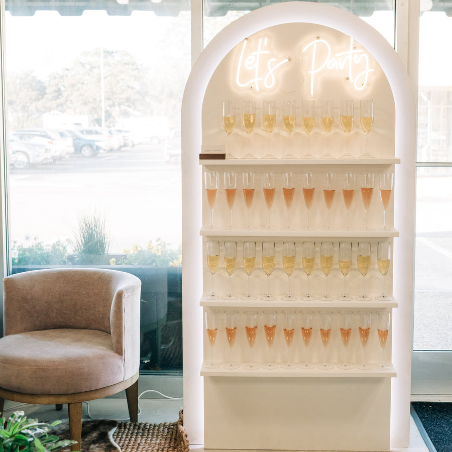 A white arched party display shelf with a glowing neon 'Let's Party' sign and multiple tiers of champagne flutes, set in a bright modern indoor space with large windows, a beige armchair, and a plant.