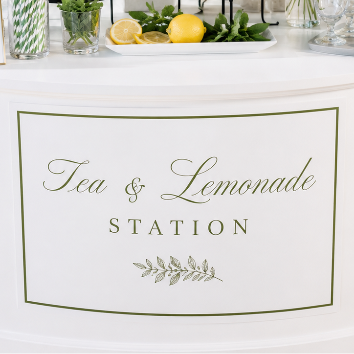 A white round beverage station table with a large sign reading 'Tea & Lemonade Station,' two glass beverage dispensers filled with iced tea and lemonade, lemon slices, flowers, straws, and clear plastic cups in a bright indoor setting.
