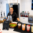Load image into Gallery viewer, A smiling woman in a black shirt stands behind a kitchen bar counter with cocktail glasses, limes, and four colorful iced drinks. A menu sign reads 'Dirty Soda Mix' and several liquor bottles are lined up on the counter.
