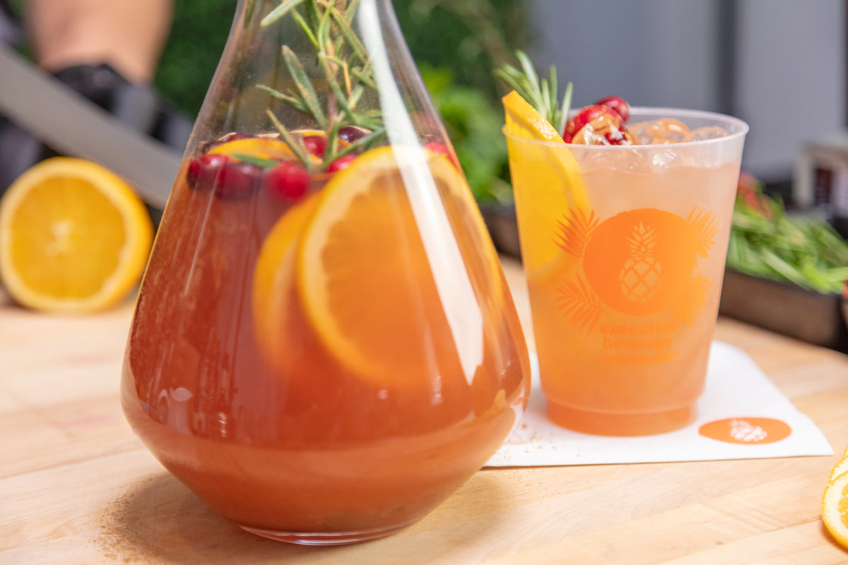 A glass beverage pitcher filled with an orange-colored drink, garnished with orange slices, cranberries, and rosemary, shown next to a matching clear plastic cup on a wooden surface.