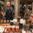 Load image into Gallery viewer, A smiling man in a black apron stands behind a hot chocolate bar setup with cups, marshmallows, candy canes, and condiments in a warmly lit indoor event space.
