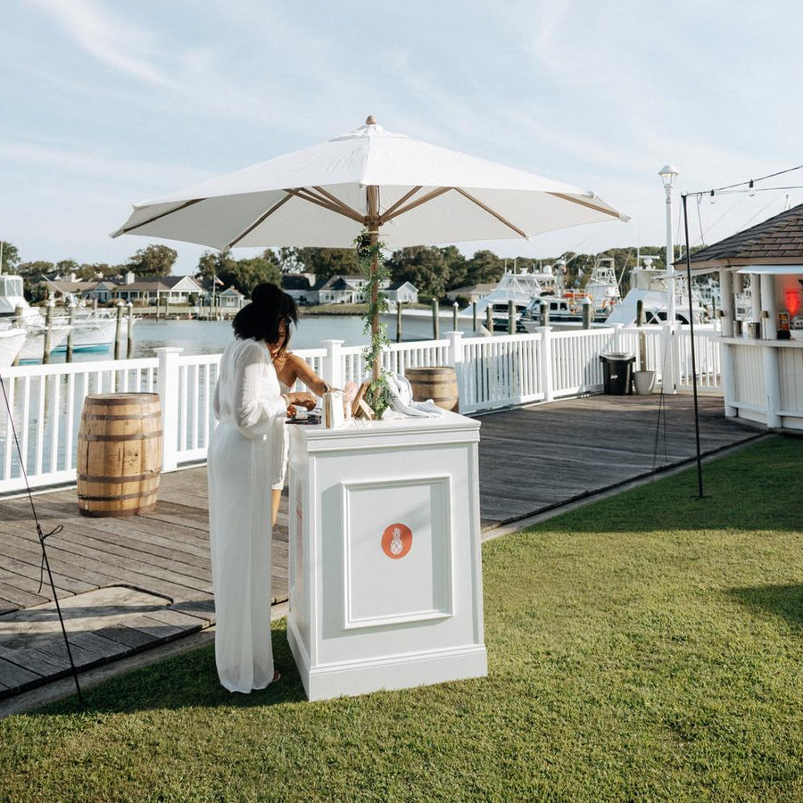A woman in a white outfit stands beside a white outdoor cocktail table box under a large white patio umbrella on a dockside lawn. The setting includes grass, a wooden dock, water, boats, and a small pavilion in the background.