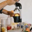 Load image into Gallery viewer, A person using a manual citrus juicer to press an apple over a glass, with bottles and cans visible in the background on a counter.

