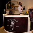 Load image into Gallery viewer, A curved bar counter with a white front and dark burgundy side panel reading 'THE GOODRICH Club CASINO NIGHT,' with a bartender in a white shirt and red bow tie standing behind it in an indoor event space.
