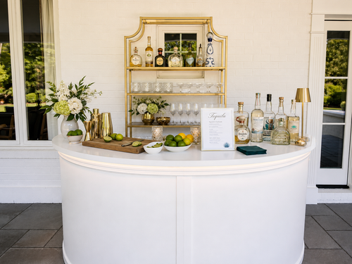 A white curved cocktail bar counter with a white top, styled with various liquor bottles, gold shakers, limes, flowers, and a gold shelving unit in the background.