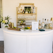 Load image into Gallery viewer, A white curved cocktail bar counter with a white top, styled with various liquor bottles, gold shakers, limes, flowers, and a gold shelving unit in the background.
