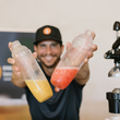 Load image into Gallery viewer, A man wearing a black cap and dark shirt holds up two clear plastic bottles containing yellow and pink-orange liquids, with a black manual juicer visible on the right side of the frame.
