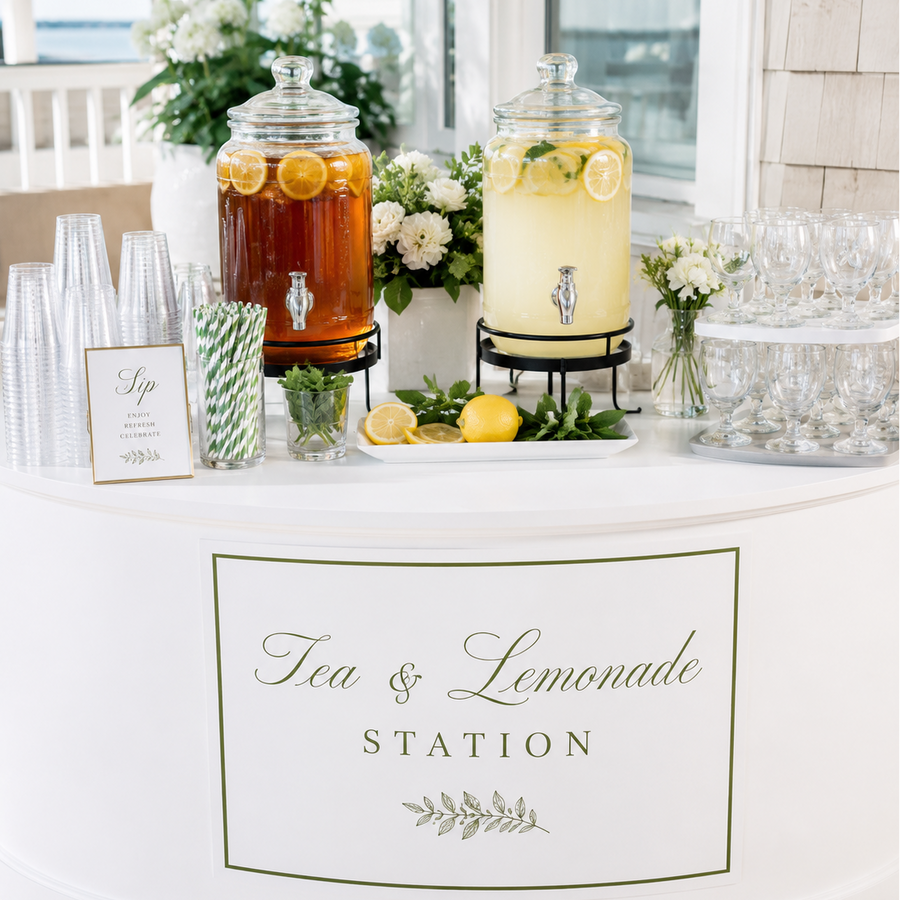 A white round beverage station table with a large sign reading 'Tea & Lemonade Station,' two glass beverage dispensers filled with iced tea and lemonade, lemon slices, flowers, straws, and clear plastic cups in a bright indoor setting.