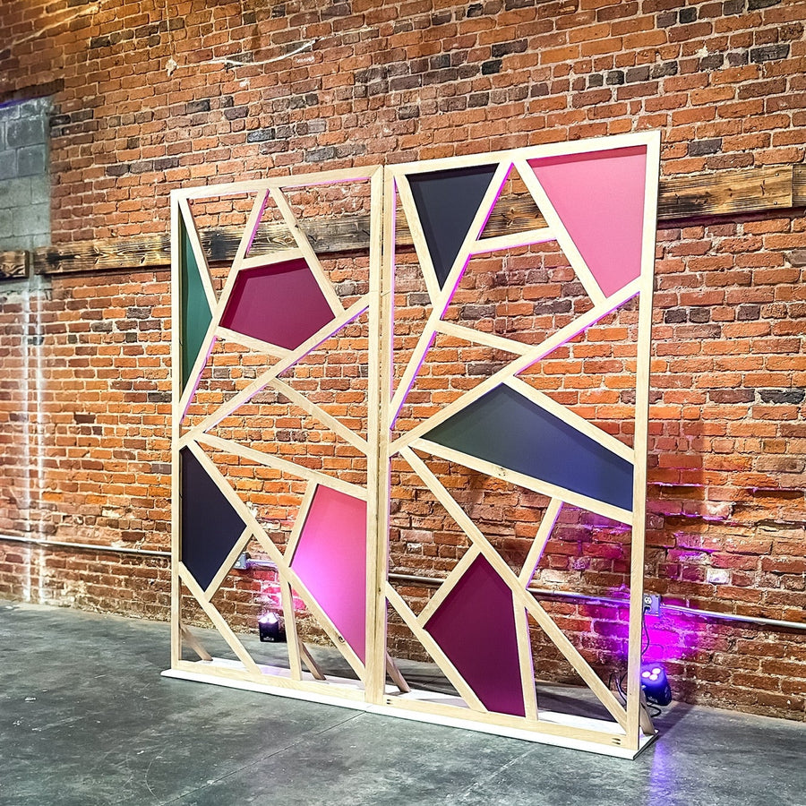 A modern geometric wooden screen panel with colorful triangular panels in pink, purple, and black, set against a brick wall in an industrial loft space.