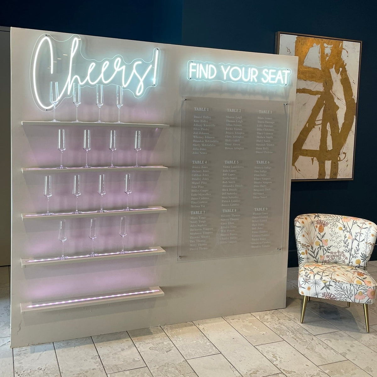 A large event seating and drink display wall with illuminated 'Cheers!' lettering, clear glass holders on shelves, a seating chart, and a patterned accent chair in a modern indoor space with dark blue walls and stone tile flooring.