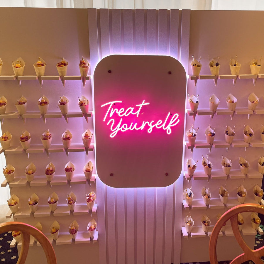 A pink neon sign reading 'Treat Yourself' mounted on a white slatted display wall filled with many small cone desserts, with chairs partially visible in the foreground.