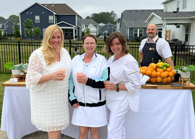 An Executive Beverage bartender with hosts at an all-white backyard party, behind a linen-draped bar table set with a fresh citrus and herb display.