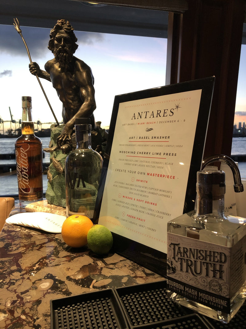 A custom Art Basel Miami Beach cocktail menu on the Antares yacht bar, framed beside bourbon and fresh citrus.
