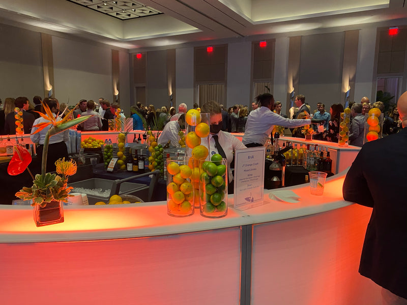 Bartenders working a busy curved LED bar at a corporate event, guests lined up along the rail beside towers of fresh citrus.