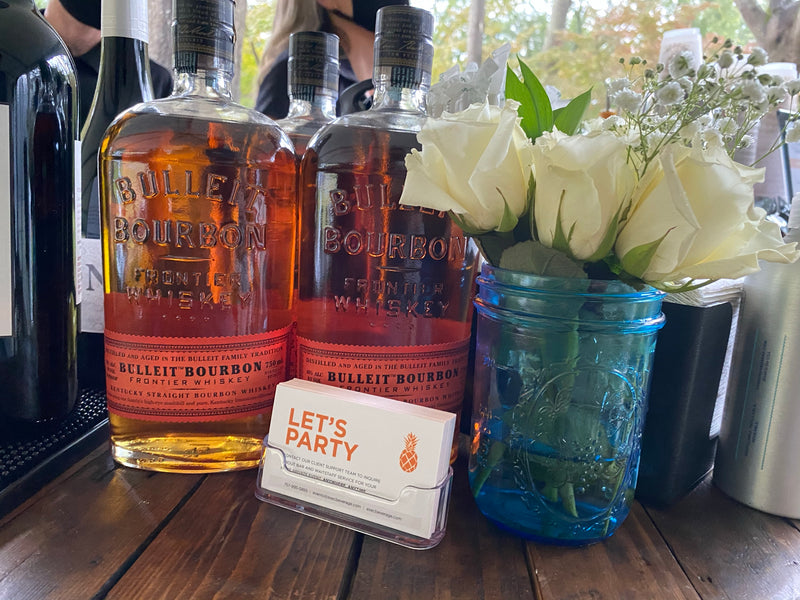 A close-up of an Executive Beverage bar top: bourbon bottles beside white roses in a blue mason jar and a Let’s Party card.