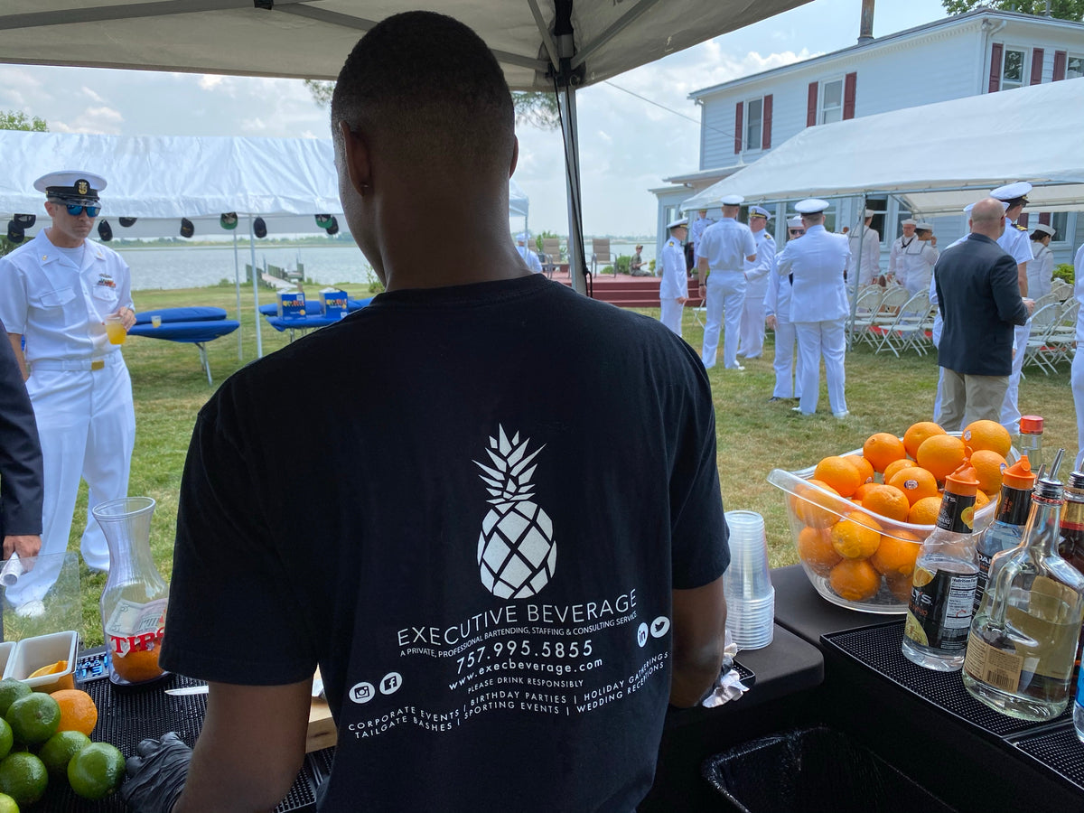 An Executive Beverage bartender working an outdoor bar at a Navy event, sailors in dress whites on the lawn beyond the tent.