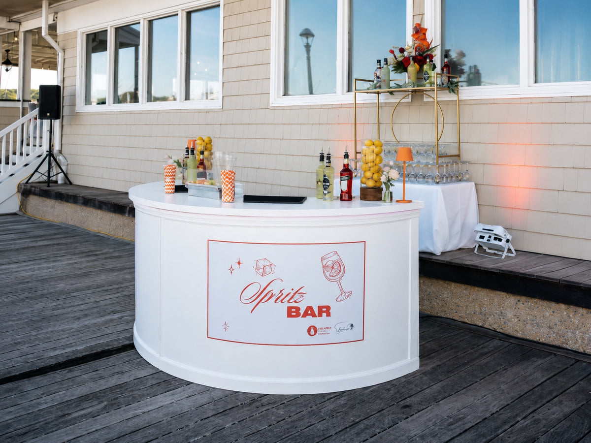 A curved white event bar dressed as a Spritz Bar with custom signage, set up on the deck of a waterfront club with a gold bar cart and glassware behind it.