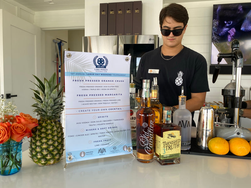 An Executive Beverage bartender beside a printed menu of fresh-pressed Orange Crush and fresh-pressed margaritas, with pineapple, roses and spirits staged along the bar.