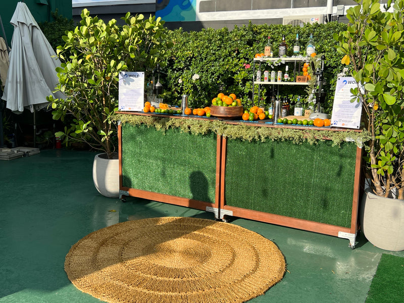A garden hedge bar at a Miami event with a full spirits display and fresh fruit along the rail.