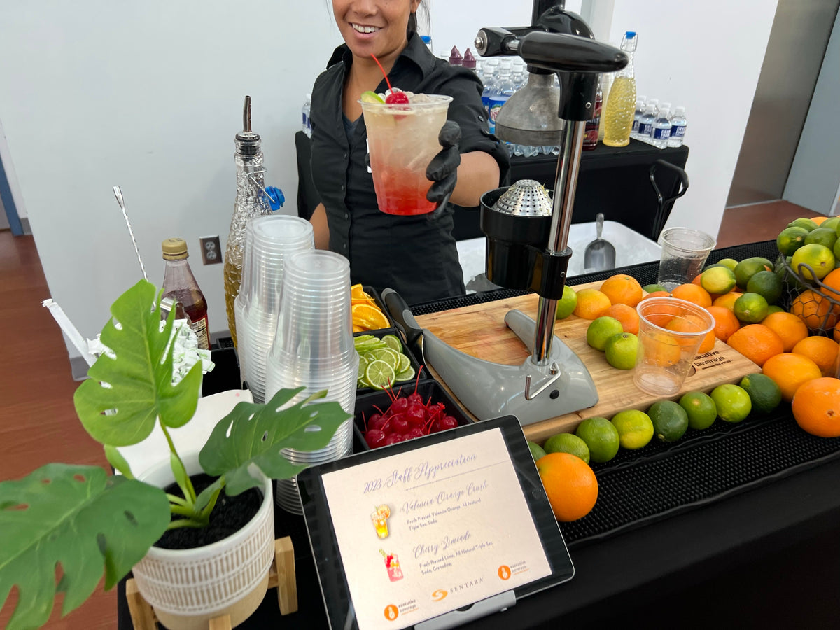 An Executive Beverage bartender at a Sentara Doctors’ Day appreciation event in Newport News, holding up a fresh-pressed mocktail beside a citrus press and crates of oranges and limes.