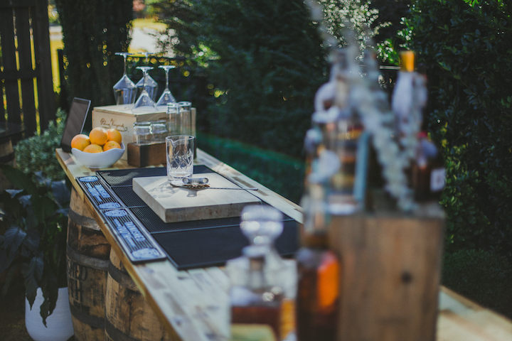 A whiskey barrel bar rental with a reclaimed bourbon barrel base and plank top, set with glassware, bourbon and a bowl of oranges at an outdoor event.