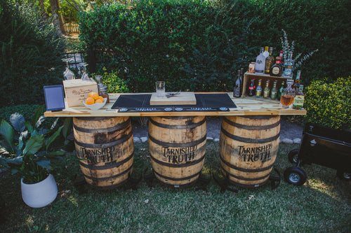 The Triple-Barrel Bar set up on a lawn, three reclaimed bourbon barrels supporting a long plank bar top with spirits, glassware and a bowl of oranges.