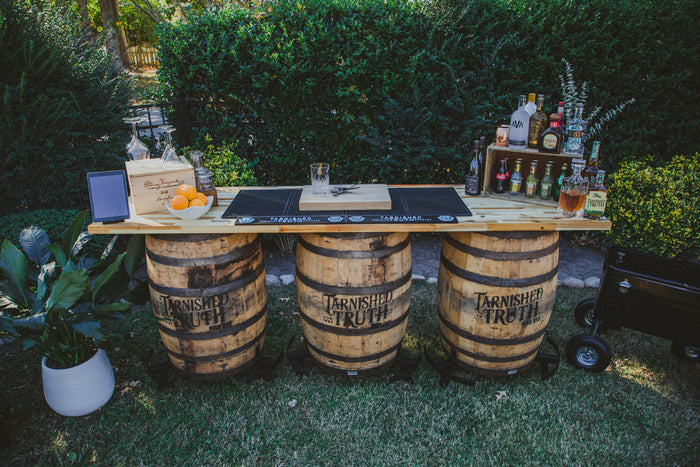The Triple-Barrel Bar set up on a lawn, three reclaimed bourbon barrels supporting a long plank bar top with spirits, glassware and a bowl of oranges.