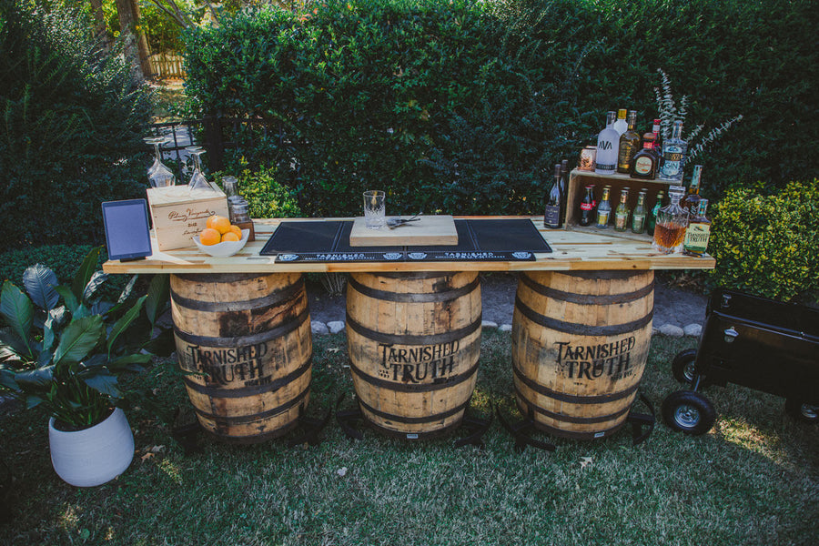 The Triple-Barrel Bar set up on a lawn, three reclaimed bourbon barrels supporting a long plank bar top with spirits, glassware and a bowl of oranges.