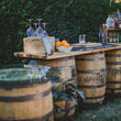 Load image into Gallery viewer, The Total Barrel Package set up outdoors, whiskey barrels carrying a plank bar top with spirits, glassware and a bowl of oranges.
